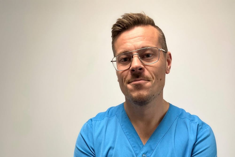 Porträtt av en person i vårdkläder mot neutral bakgrund.