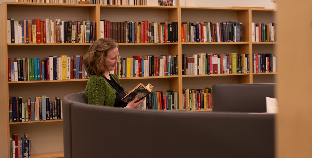 En kvinna i grön tröja sitter i en grå fåtölj i ett bibliotek och läser en bok, med bokhyllor fyllda av böcker i bakgrunden.