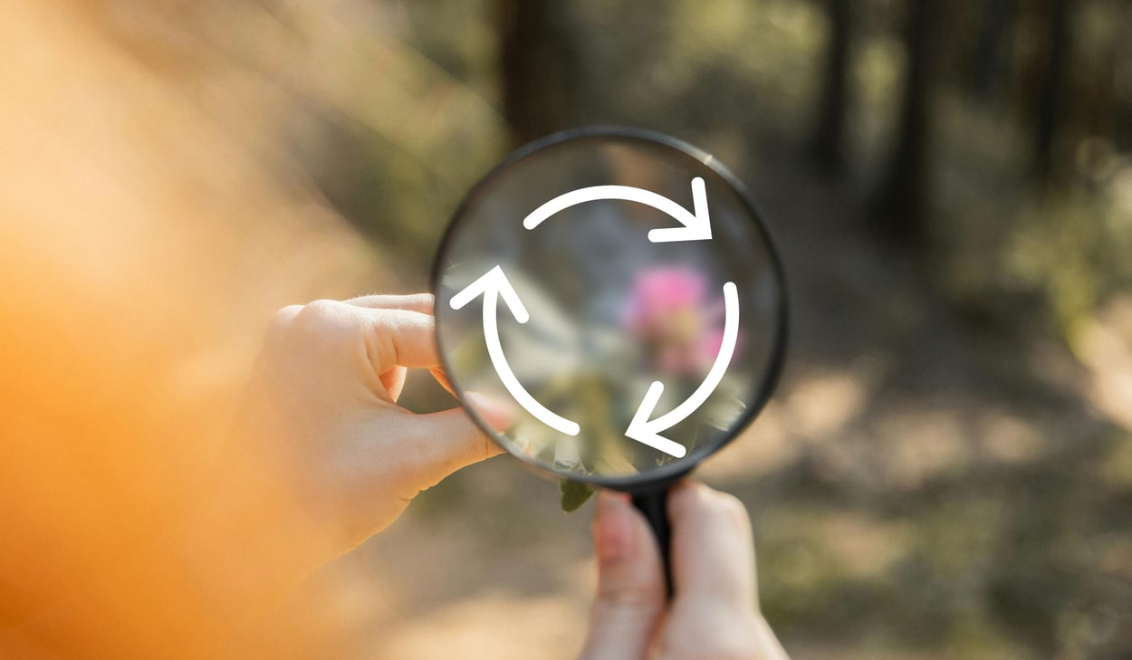 Hand som håller i ett förstoringsglas med skog i bakgrunden. I förstoringsglaset syns en illustration av cirkulär ekonomi