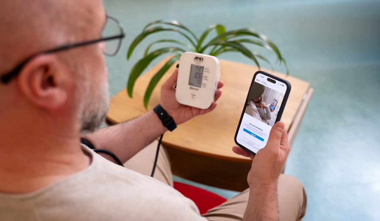 En person sitter ner och mäter sitt blodtryck med en digital blodtrycksmätare. Personen håller mätaren i ena handen och en mobiltelefon i den andra, där en app med vårdrelaterat innehåll visas. 
