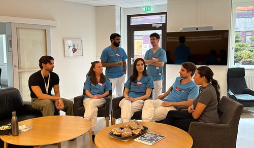 Samlingslokal med vita väggar, två bruna bord, sju personer som sitter och samtalar. Fyra personer i blåa och vita kläder och två personer i privata kläder. AT/BT centrum